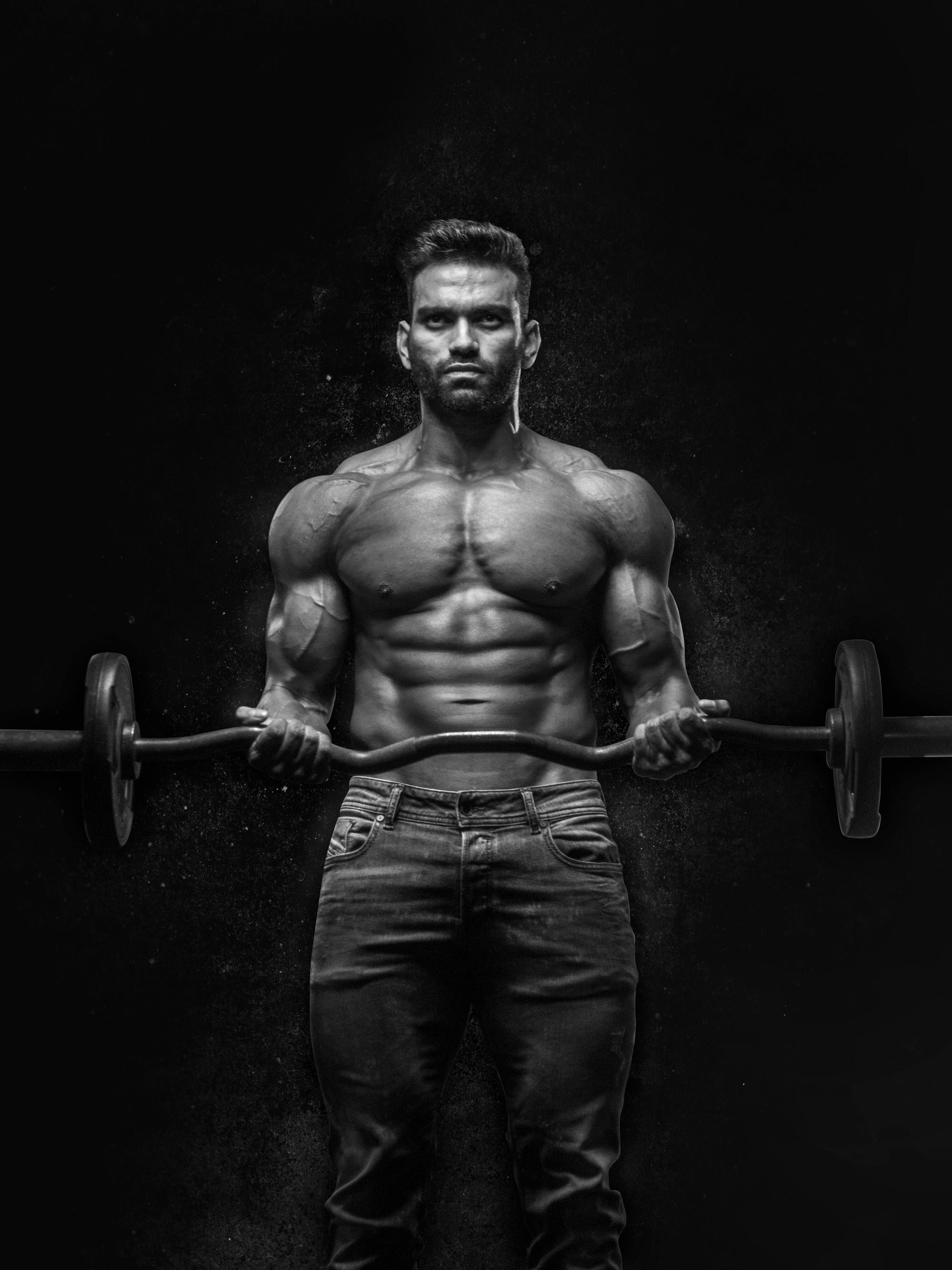 A black and white image of a muscular man lifting a barbell. The man is shirtless, showcasing his defined abs and upper body muscles, and is wearing jeans. He is standing against a dark background, looking directly at the camera with a serious expression.